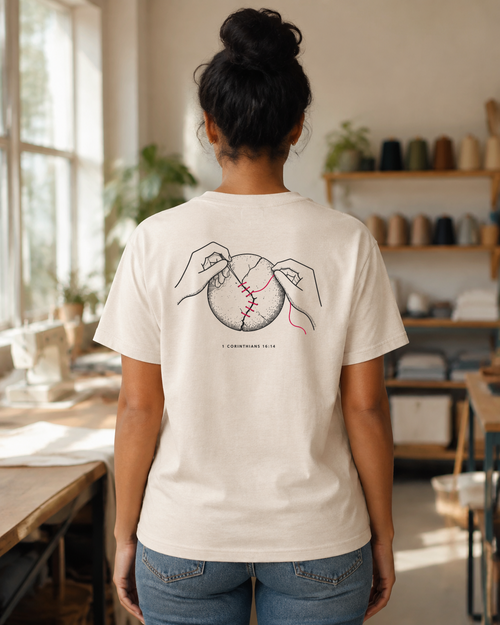 Back of a cream tee with hands stitching a cracked circle using red thread and 1 Corinthians 16:14. Back print shown.